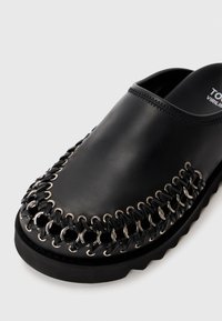 Chaussure noire en cuir à enfiler avec un dos ouvert, décorée d'œillets en argent et de lacets tressés noirs autour des orteils et des côtés.