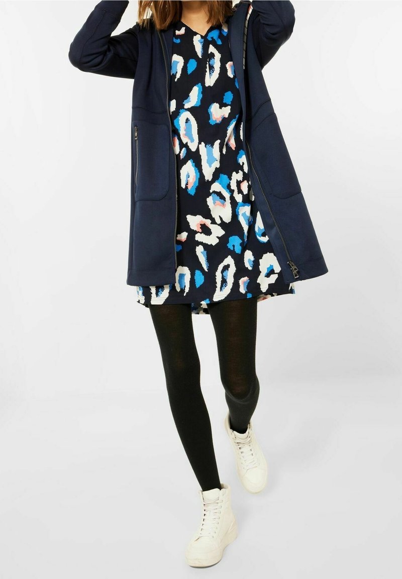 Femme portant un sweat à capuche bleu marine sur une robe noire avec un imprimé abstrait bleu, blanc et rose, des collants noirs et des baskets blanches, marchant vers l'avant.