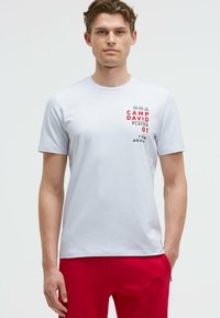 White cotton t-shirt with short sleeves, featuring red and black graphic text and symbols on the front. Crew neckline, casual fit.