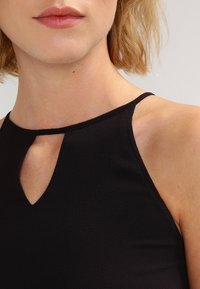 Musta hihaton toppi, jossa on avainreiän kaulus, kapeat hihnat ja pehmeä, tekstuuriainen kangas. Ei näkyviä lisäkuvioita tai varusteita.