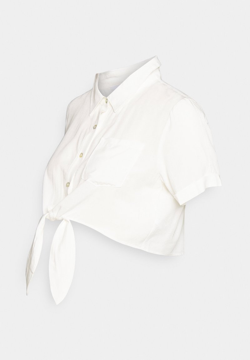 Chemise blanche à manches courtes boutonnée, raccourcie et nouée devant avec une poche poitrine sur fond uni.