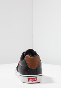 Musta lenkkari, jossa on teksturoitu päällinen, ruskea nahkainen kantakappaleen korostus, valkoinen kumipohja ja punainen Levi's-logo sivulla.