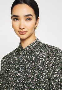 Camicia nera a fiori con piccoli fiori bianchi e verdi, colletto, chiusura frontale con bottoni e tessuto testurizzato con una finitura morbida.