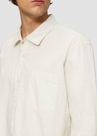 Chemise longue beige clair à manches longues en tissu doux, dotée d'un devant boutonné, d'un col rabattu et d'une poche sur la poitrine côté gauche.