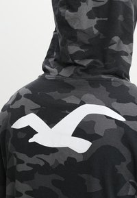 Svart och grå camouflage hoodie med en stor vit mås grafik på baksidan. Mjuk tyg med en texturerad yta.