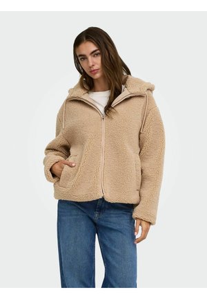 Femme portant une veste sherpa polaire beige zippée avec capuche et un jean bleu, debout avec une main dans la poche devant un fond uni.