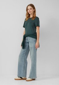 T-shirt à manches courtes vert foncé, associé à un jean large bleu clair et des baskets crème, avec un pull en maille verte noué autour de la taille.