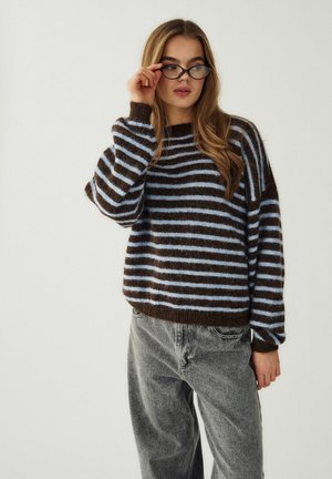 Fiatal nő, amint fekete, macskaszem alakú szemüveget igazít, barna és világoskék csíkos pulóvert és fakó szürke farmert visel, egyszerű háttér előtt áll.