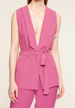 Różowa, bezrękawnikowa, dopasowana kamizelka z głębokim dekoltem w serek i pasem wiązanym w talii, noszona z różowymi spodniami na osobie z tatuażem na ramieniu.