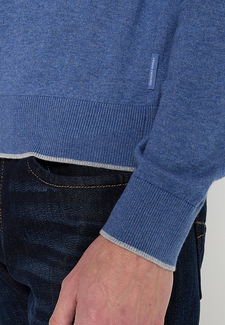Blauer gestrickter Pullover mit gerippten Bündchen und Saum, der am unteren Rand einen grauen Akzent und ein kleines Markenetikett an der Seite aufweist.