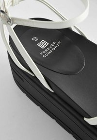 Juoda platforminė šlepetė su baltais dirželiais. Yra paminkštintas juodas padas su "FOREVER COMFORT" logotipu. Reguliuojamas sagtis ir tekstūruotas padas.