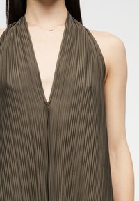 Γυναίκα που φοράει ελιά πράσινο πλισέ φόρεμα με λαιμόκοψη halter και βαθύ V-λαιμόκοψη, με λεπτό χρυσό κολιέ με μαργαριταρένιο μενταγιόν.