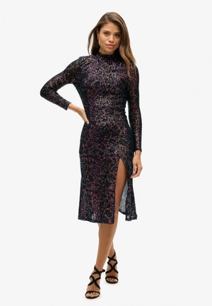 Sort pangfarvet kjole med lange ærmer, høj hals, sideslids og tætsiddende design. Matchet med sorte sandaler med stropper. Glat, glitrende tekstur.