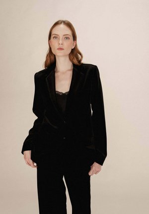 Blazer en velours noir avec un design ajusté, des revers crantés et des boutons à l'avant. Associé à un haut noir à bordure en dentelle en dessous. Texture lisse.