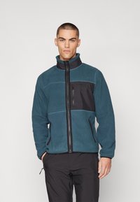 Chaqueta de forro polar en azul verdoso con un panel frontal negro. Cuenta con cremallera completa, cuello alto y bolsillos laterales. Tejido texturizado con costuras en contraste.