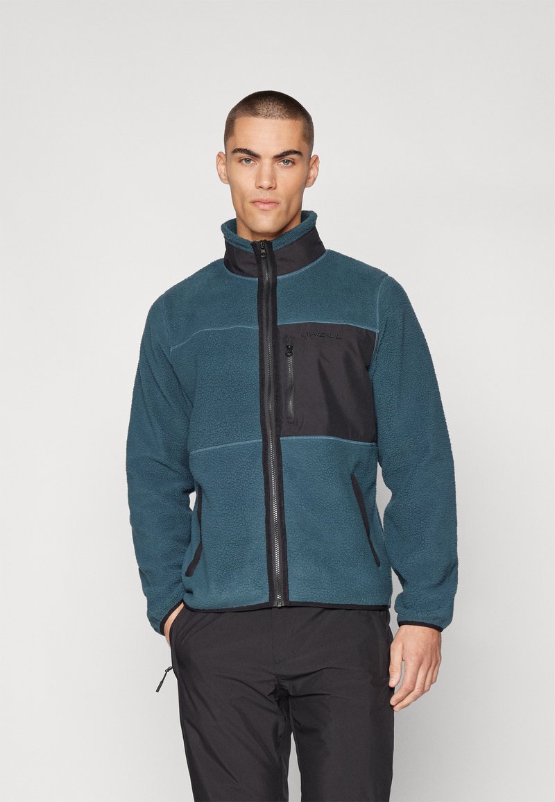 Chaqueta de forro polar en azul verdoso con un panel frontal negro. Cuenta con cremallera completa, cuello alto y bolsillos laterales. Tejido texturizado con costuras en contraste.