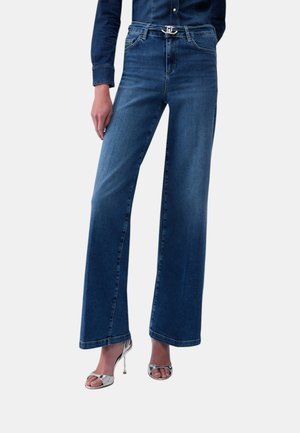 Personne portant un jean bleu à jambes larges avec une boucle de ceinture argentée et des chaussures argentées à talons hauts et à bout ouvert, debout devant un fond uni.