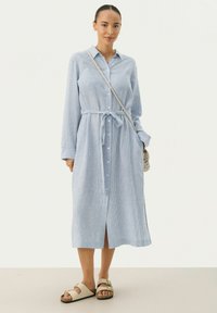 Abito lungo a righe blu chiaro con bottoni frontali, vita con cintura e maniche lunghe. Abbinato a sandali bianchi e a una borsa a tracolla.