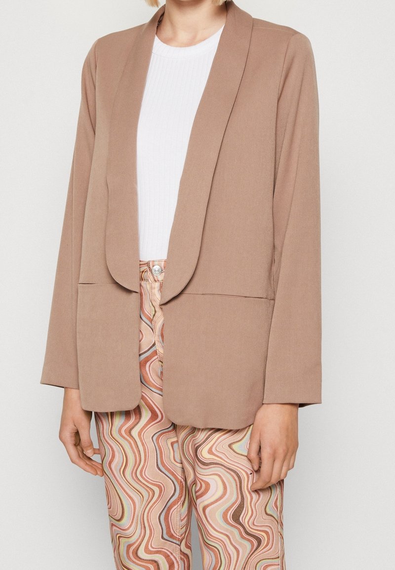 Femme portant un blazer beige sur un top blanc avec un pantalon taille haute présentant un motif ondulé multicolore dans des tons pastel doux.