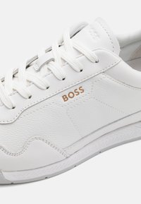Hvit lær sneakers med teksturerte paneler, med hvite snører og en bronsefarge logo på siden. Gummisåle med et mønster.