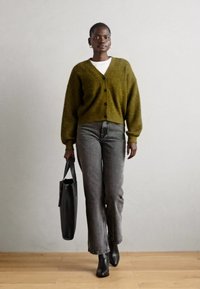 Γυναίκα που περπατάει σε εσωτερικό χώρο φορώντας ελαιόχρωμο cardigan, λευκό πουκάμισο, γκρι τζιν, μαύρες μπότες και κρατώντας μια μαύρη τσάντα tote.