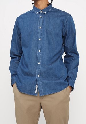Mann trägt ein blaues Jeanshemd mit langen Ärmeln und beige Hose, Hände teilweise in den Taschen, vor einem einfarbigen Hintergrund.