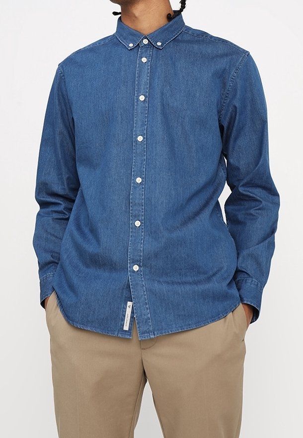 Homme portant une chemise en jean bleue à manches longues boutonnée avec un pantalon beige, les mains partiellement dans les poches, sur un fond uni.