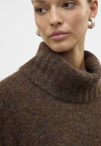 Pull en laine marron à col roulé avec un tricot texturé, de subtils éclats colorés et un col haut. Grandes boucles d'oreilles circulaires en argent visibles.