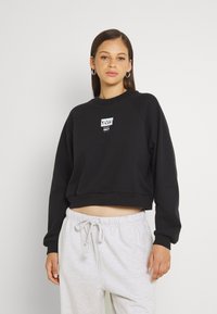 Felpa nera corta con maniche raglan, caratterizzata da una toppa con logo frontale. Abbinata a pantaloni della tuta grigi chiari, adatta per l'abbigliamento da casa.
