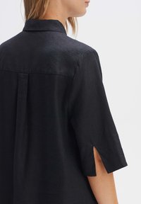 Camicia in lino nera con colletto, maniche corte con fessure laterali e dettaglio a plissettatura sul retro, che mette in risalto un tessuto testurizzato.