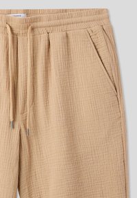 Pantaloni beige texturizzati con tessuto stropicciato, vita elasticizzata, chiusura con cordoncino e tasche laterali.