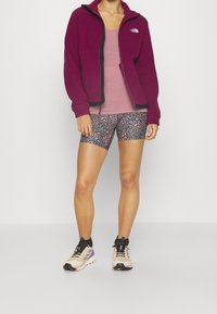 Lila fleecetröja, ljusrosa linne, mönstrade shorts och beigea sportskor. Funktioner inkluderar en dragkedja fram och svarta accenter.