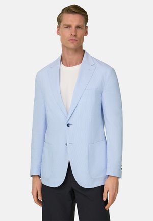 Man draagt lichtblauwe getextureerde blazer, wit shirt met ronde hals en zwarte broek, staand tegen een effen achtergrond.