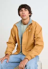 Giacca leggera giallo-marrone con interno blu-grigio, chiusura con zip e polsini elastici. Presenta un piccolo logo sul petto.