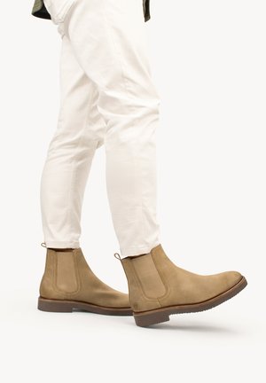 Botas Chelsea de ante marrón con paneles laterales elásticos, tiradores y suelas de goma, combinadas con pantalones ajustados blancos.