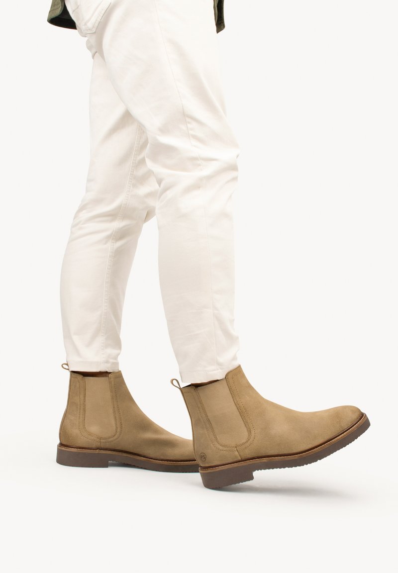 Botas Chelsea de ante marrón con paneles laterales elásticos, tiradores y suelas de goma, combinadas con pantalones ajustados blancos.