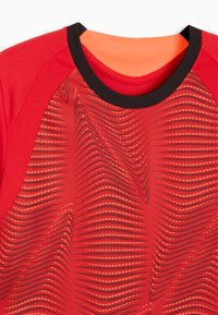 Camiseta deportiva roja con un patrón de ondas en rojo oscuro y naranja. Mangas cortas, cuello negro y textura de tela suave.