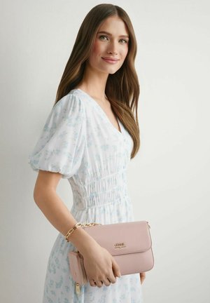 Młoda kobieta w jasnoniebieskiej sukience w kwiaty, trzymająca bladoróżową portmonetkę z złotą bransoletką na nadgarstku.