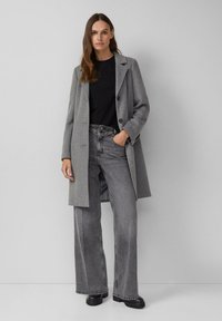 Grijze wollen jas met inkepingen, zwart shirt en wijde grijze denim broek. Zwarte chunky laarzen en palmzakdetail op de broek.