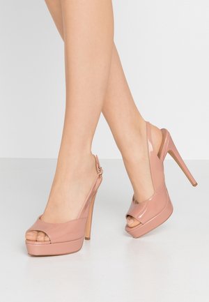 Jambes portant des sandales slingback roses blush brillantes à talons hauts avec orteils découverts et semelles compensées sur une surface lisse.