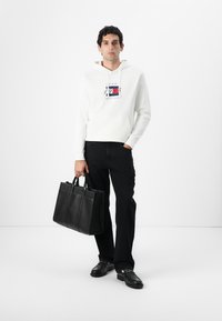 Weißer Hoodie mit Grafikdesign vorne, schwarze Hose und eine schwarze Lederhandtasche mit zwei Tragen, vor einem schlichten Hintergrund.