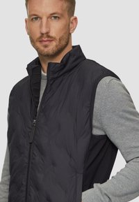 Zwarte gewatteerde vest met hoge kraag en ritssluiting. Heeft een mouwloos ontwerp over een grijze lange mouwen shirt. Gladde textuur.