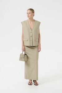 Gilet senza maniche in lino beige con due tasche frontali, abbinato a una gonna maxi coordinata; tiene una piccola borsa a tracolla con motivo a serpente.
