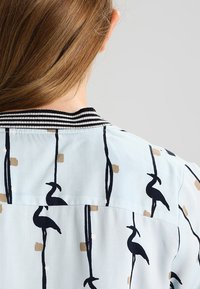 Ljust blå blus med randig krage och svarta fågelmönster. Har subtila bruna geometriska former som skapar en texturerad yta.