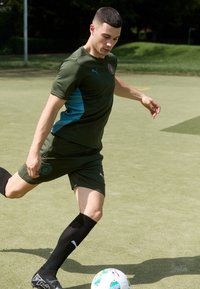 Sportists zaļā Puma krekla ar tirkīza akcentiem un melnām šortiem, melnās zeķēs, sitot futbolu uz zāles lauka.