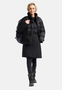 Zwarte pufferjack met een boxy pasvorm, gecombineerd met een lange zwarte jas, leggings en stijlvolle winterlaarzen met bontvoering. Rugzak inbegrepen.