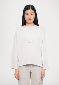 Ung kvinde med langt, mørkt hår iført en løs, lys beige sweater med brede ærmer og matchende lyse bukser.