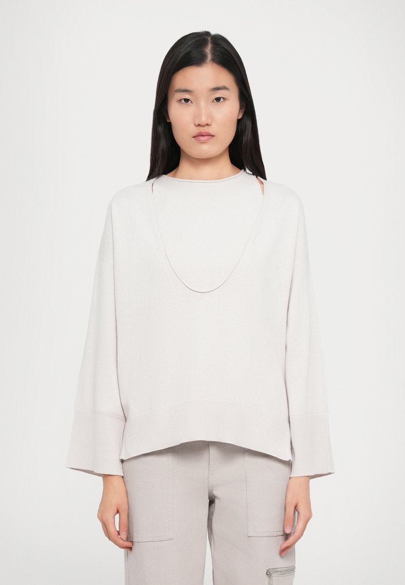 Ung kvinde med langt, mørkt hår iført en løs, lys beige sweater med brede ærmer og matchende lyse bukser.