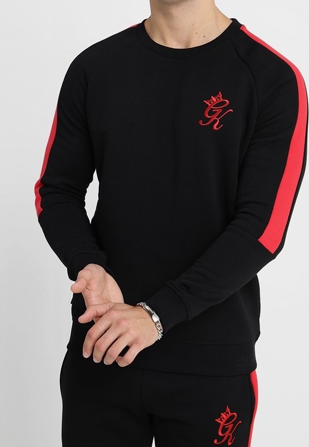 Sweatshirt noir avec des bandes latérales rouges, arborant un petit logo rouge sur la poitrine. Poignets et ourlet côtelés avec une texture de tissu lisse.