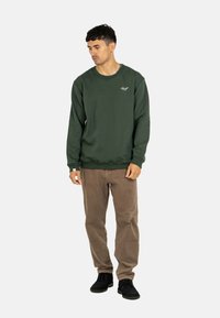 Olive green sweatshirt with ribbed cuffs and hem, featuring a small white logo on the chest, paired with light brown corduroy pants and black shoes.